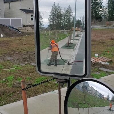Worker with pressure washer cleaning sidewalk, reflection in vehicle mirror.