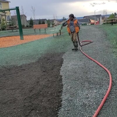 Worker spraying grass seed in a park with playground equipment in background.