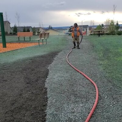 Man working with hose on landscaped park grounds near playground and benches.