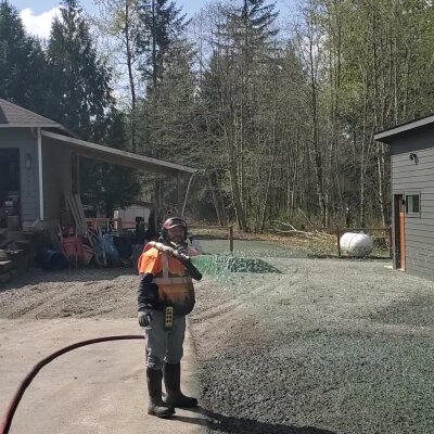 Worker spraying water on gravel near wooded area and buildings.