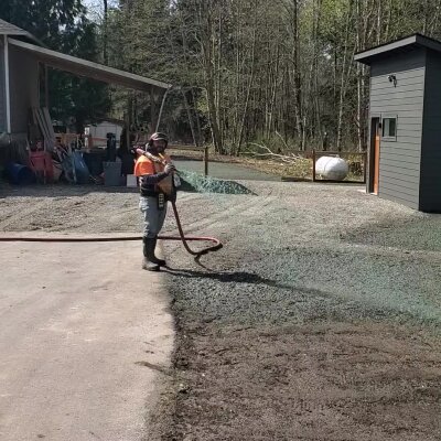 Worker using hose to apply liquid on gravel outdoors.