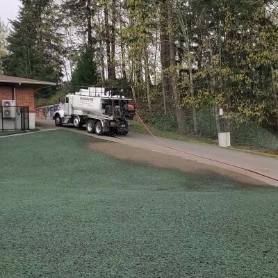 Truck spraying green liquid on grass near trees and a building.