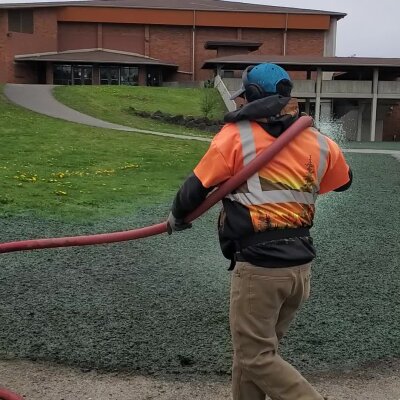 Worker with hose applying material in garden near building.