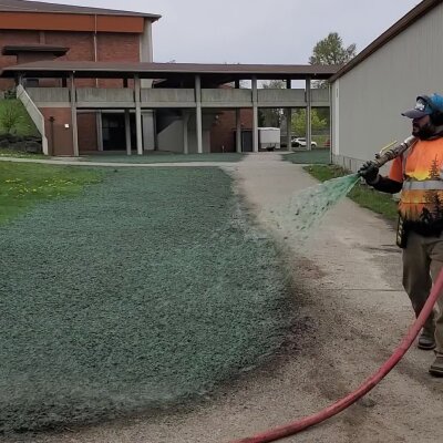Worker spraying green hydroseed mixture on soil near building.