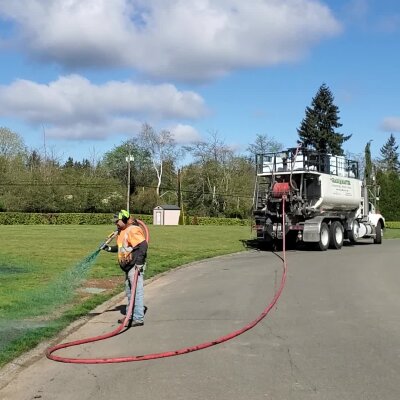 Worker spraying liquid from hose connected to a tanker truck in park.