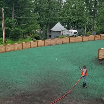 Worker hydroseeding lawn with green mulch near wooden fence.